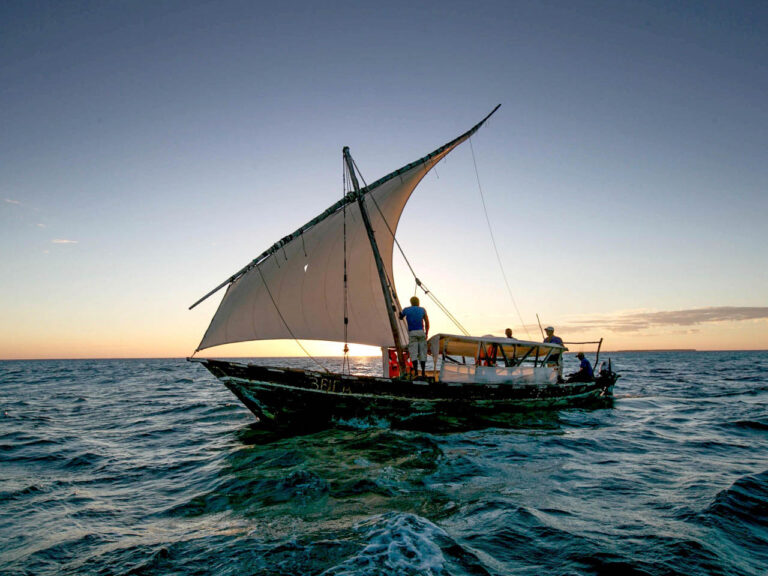 dhow met mast en man daarnaast met geheven zeil en overdekt zitgedeelte met een stuurman daarachter varend op de indische oceaan bij Fanjove