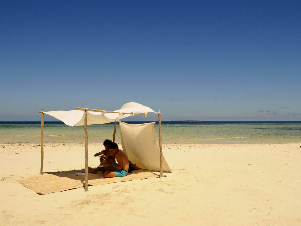 op het strand twee mensen zittend onder een een luifel bestaande uit vier houten palen met daartussen een doek gespannen uitkijkend over de indische oceaan