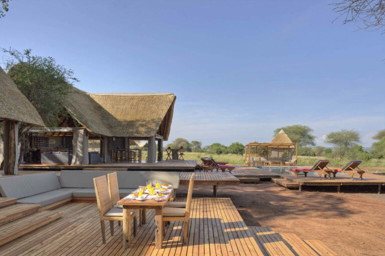 lodge bij tarangire met op de voorgrond een vlonder met daarop een gedekte tafel met stoelen daarachter een l vorige loungebank en rechts daarvan een zwembad met dekchairs