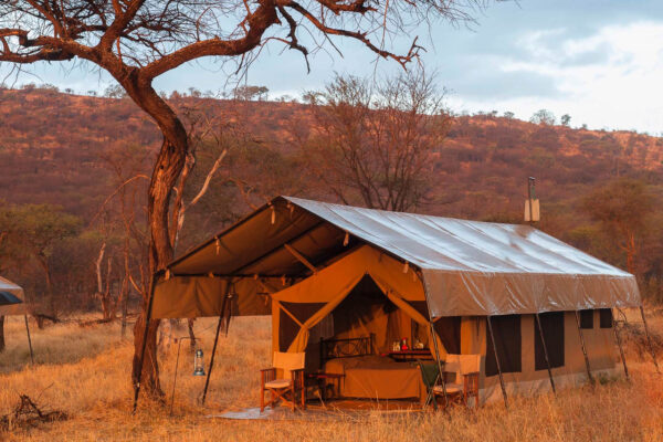 inkijk in safari tent met veranda en open geslagen deurflappen met daarbinnen een bed dit alles met een oranjegloed door de zonsondergang