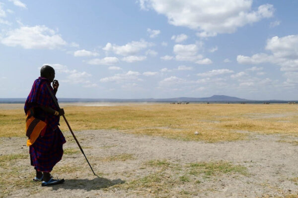 Masai man in traditionele kleding uitkijkend over een droge vlakte met aan de horizon een paar zebra's