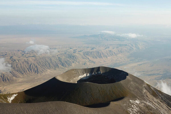 vanuit de lucht de zwarte ronde krater rand van de Ol-doinyo Lengai vulkaan met op de achtergrond een heuvelachtig landschap ontstaan door lava