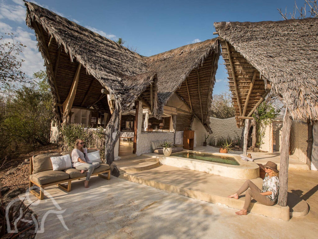 twee mensen zittend waarvan 1 op een bank en de ander tegen paal op de grond bij een bar en vijver behorend bij een luxe accomodatie in het zuider van tanzania