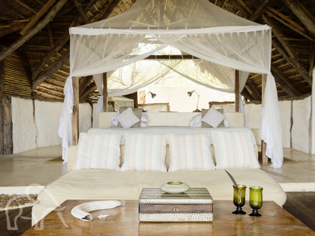 luxe grote kamer met een witte bank met kussens en daarachter een heel groot bed met klamboe omgeven door witte muren en een hoog rieten dak in zuid tanzania