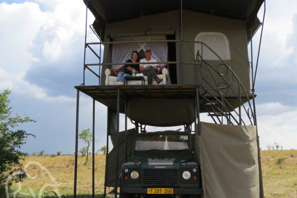 voorkant van een landrovermet recht een douche afgeschermd met canvas een trappetje naar boven waar twee mensen zitten op een bank die staat op een veranda