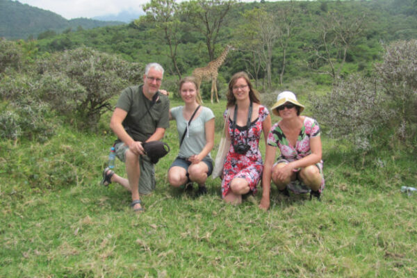 Familiefoto met een giraf op de achtergrond tijdens een wandel safari in Arusha National Park