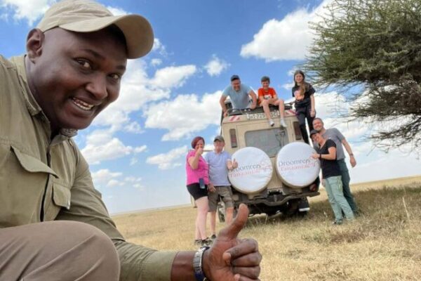 gids en op de achtergrond 7 reizigers die op safari zijn en voor op en in de safari auto staan