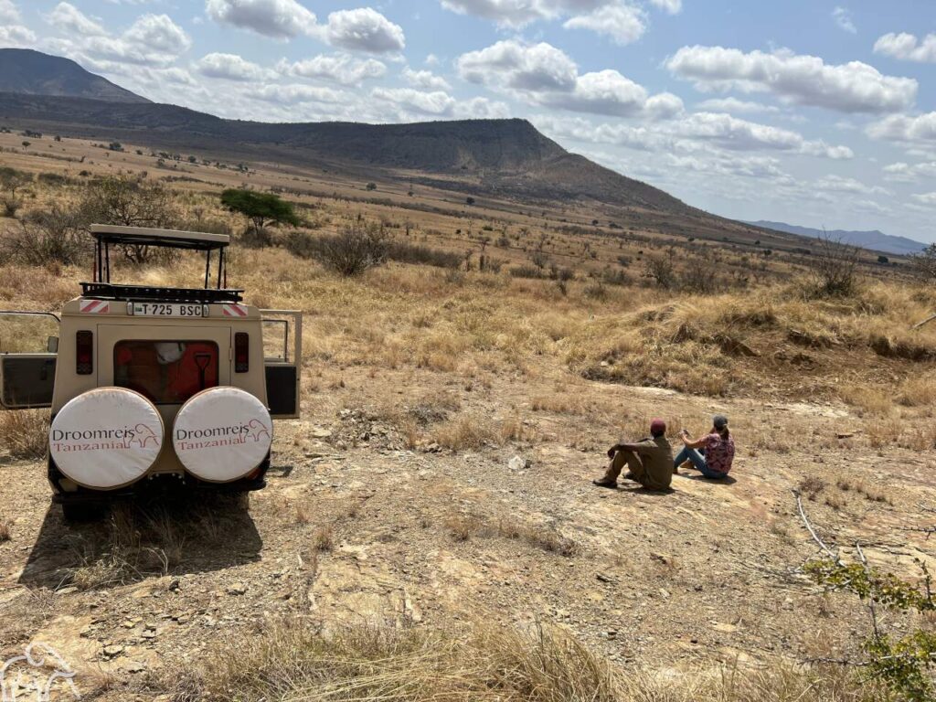 Over ons.Safari Tanzania. In Mkomazi National Park zit Wilma eigenaar van Droomreis Tanzania samen met de gids op een steen met verderop de safariauto van Droomreis Tanzania. Ze kijken uit over goudgele vlaktes en bergen. Een adembenemend landschap