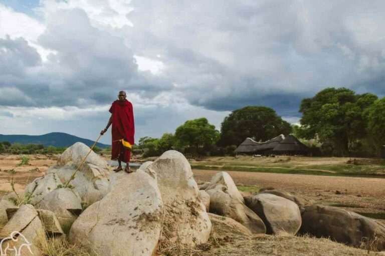 Masai man op grote rotsen met daarachter de Ruaha rivier en een accommodatie in Ruaha National Park Tanzania