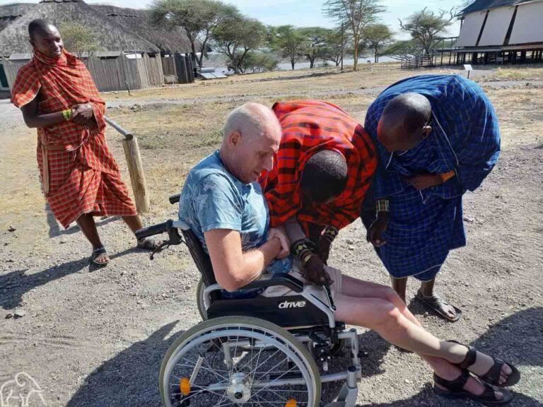 Drie Masai mannen waarvan twee voorovergebogen bij een man in een rolstoel