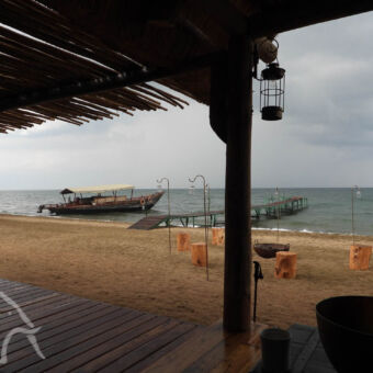 uitzicht Mbali Mbali camp uitzicht vanaf de veranda van Mbali Mbali camp op het strand met krukjes van hout een steiger in het Tanganyika meer en een oude vissersboot