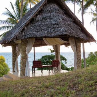Op vier pilaren rustend puntdak van riet met daaronder twee en een tafeltje met vandaar uitzicht op het strand palmbomen en de indische oceaan op Mafia eiland