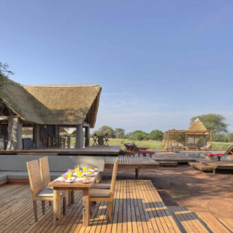 lodge bij tarangire met op de voorgrond een vlonder met daarop een gedekte tafel met stoelen daarachter een l vorige loungebank en rechts daarvan een zwembad met dekchairs