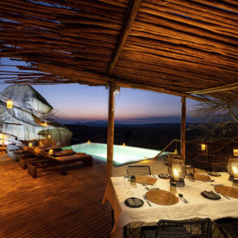 ruaha bi nacht bij een lodge een wit gedekte tafel met kaarsjes op een terras onder een afdak met daarachter een verlicht zwembad