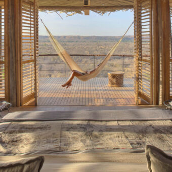 grote kamer in lodge waarbij we kijken van achter het bed en kijken over de houten veranda over ruaha national park. tussen de openslaande deuren hangt een hangmat en links en recht van de deuren staan rieten stoelen