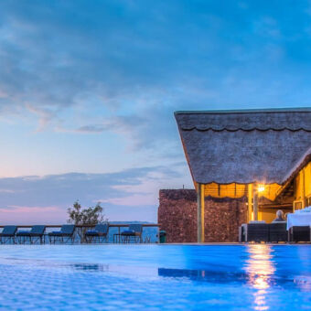 uitzicht met camera op de grond uitkijken over een zwembad met daarachter een lounge ruimte met veel glas en een rietendak