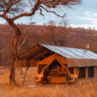 comfortabel inkijk in safari tent met veranda en open geslagen deurflappen met daarbinnen een bed dit alles met een oranjegloed door de zonsondergang