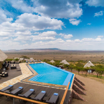luxe tented lodge in de Serengeti luxe lodge met links lounge banken op de veranda en in het midden een groot zwembad met ligstoelen en daarachter rietgedekte huisje die uitkijken over de vlaktes van de serengeti
