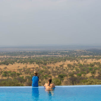 zwembad bij een lodge met jongetje zittend op de rand en vrouw naast hem in het zwembad beide kijkend over de serengeti