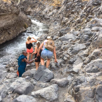 waterfall hike drie vrouwen en een gids op excursie lopend over en tussen de rotsen door naast een riviertje op weg naar een waterval bij Lake Natron