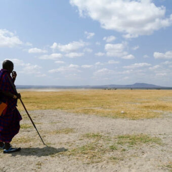 Masai man Masai man in traditionele kleding uitkijkend over een droge vlakte met aan de horizon een paar zebra's