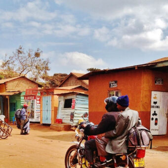 straatbeeld in karate rijdt een brommer met twee mensen op een onverharde weg met daarvoor een jongen met een handkar en een man met heel veel spullen op z'n rug
