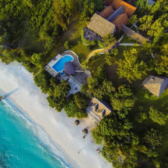 lodge van bovenaf vanuit de lucht een uitzicht op het Manta resort met een klein stukje zee wit strand een zwembad het hoofdgebouw met rieten dak en kamers aan zee
