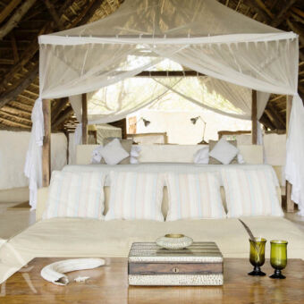 luxe grote kamer met een witte bank met kussens en daarachter een heel groot bed met klamboe omgeven door witte muren en een hoog rieten dak in zuid tanzania