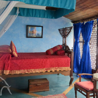 kleurrijke kamers kamer van het Emerson Spice hotel met traditioneel bed van hout en gedraaide poten met een rood dekbed blauwe gordijnen voor de ramen en een grote klamboe