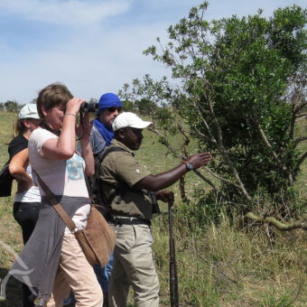 de ogen van de gids zijn beter een meerdaagse wandelsafari waarbij een vrouw met verrekijker en gids die wijst en man die luistert op de vlakte van de serengeti