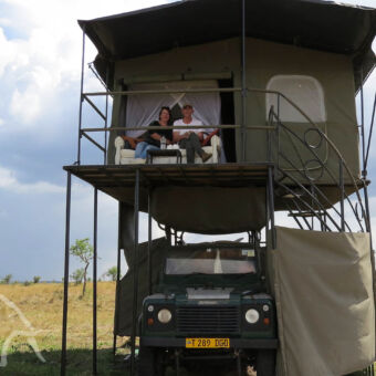 genieten van het uitzicht voorkant van een landrovermet recht een douche afgeschermd met canvas een trappetje naar boven waar twee mensen zitten op een bank die staat op een veranda