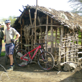 man en gids staand bij een mountainbike en houtenhutje klaar om op bezoek te gaan bij mensen in het dorp