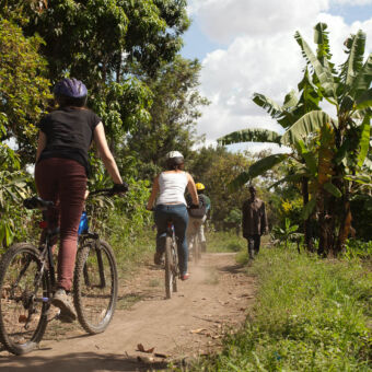 over de paadjes 3 mensen fietsend op een mountainbike waarvan een gids op een onverharde weg met bananenbomen recht en een wandelende tanzaniaanse aan de kant