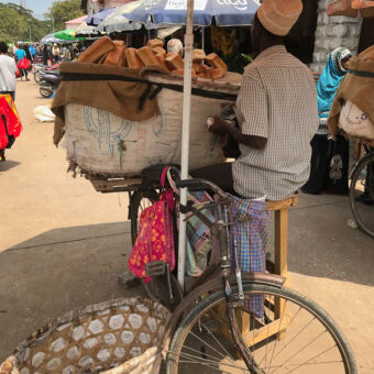 man zittend op een houten krukje naast zijn fiets die volgepakt is met een hele grote zak vol met broden op een markt in tanzania