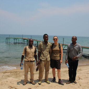 Vier mensen waarvan 1 vrouw op het strand van lake Tanganyika met op de achtergrond het meer en een steiger mahale tanzania
