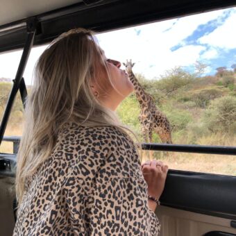 Leuke foto's tijdens je safari. Heb jij ooit een giraf gekust? Safari rondreizen door Tanzania ter inspiratie. Er staat een vrouw in de safariauto en op de achtergrond staat een giraf. De foto is zo gemaakt dat het lijkt of de vrouw de giraf kust.