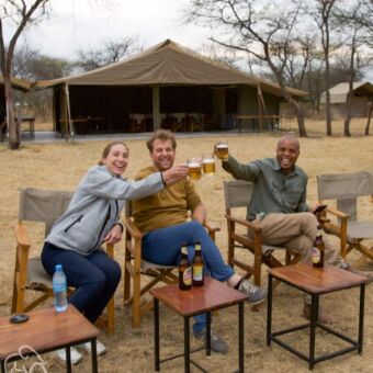 lachende man en vrouw zittend samen met hun gids proosten met een biertje bij een tented camp tanzania