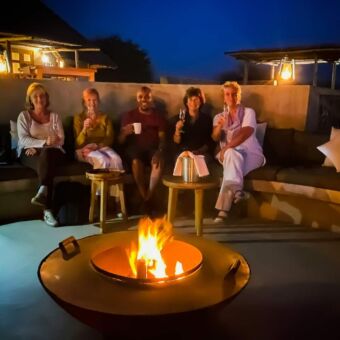 Vier vriendinnen met hun gids bij het kamper met een drankje in de prachte lodge van Manyara Secrets Safari in Tanzania. Vier vriendinnen op leeftijd zittend met hun gids in het midden bij een kampvuur met een drankje. Ze zijn bij Manyara Secrets.