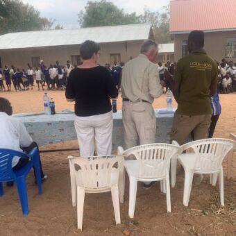 drie mensen die staan bij een tafeltje en een school bezoeken waar op de achtergrond allemaal kinderen zijn