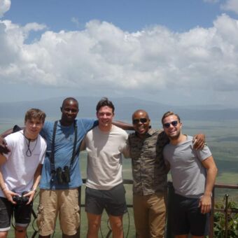 3 gasten en hun gidsen je aankijkend bij het uitzichtspunt van de nogorongoro krater. op de achtergrond zie je de groene krater