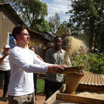 man met kom in de hand die probeert de vliesjes van het mais te scheiden door het omhoog te gooien. Een andere man kijkt toe hoe de toerist het doet