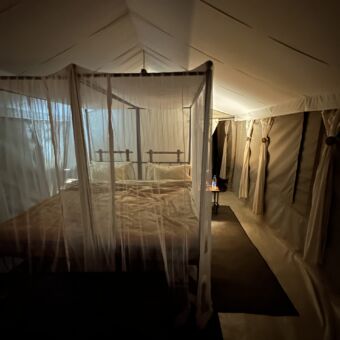 je kijkt door het gaas in de safaritent waar een groot bed staat mooi opgemaakt met witte lakens.