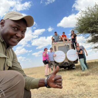gids en op de achtergrond 7 reizigers die op safari zijn en voor op en in de safari auto staan