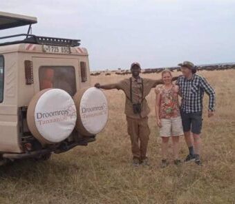 op de voorgrond de safariauto van droomreis tanzania met daarnaast de gids en twee gasten tijdens hun huwelijksreis. op de achtergrond to aan de horizon een grote groep gnoes tijdens de grote migratie op de serengeti
