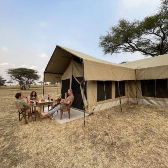 Op de Serengeti heerlijk borrelen voor je safari tent. Houd je camera bij de hand er kan zomaar een dier voorbij komen. Serengeti rondreizen in Tanzania ter inspiratie. Op de vlaktes van de Serengeti staat een safari tent waarvoor 3 mensen zitten te borrelen.