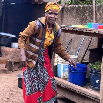 Tanzaniaanse vrouw met hoofddoek en kleurrijke kleding staat voor een washok met emmers. Ze kijkt je half lachend aan