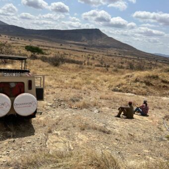 Over ons.Safari Tanzania. In Mkomazi National Park zit Wilma eigenaar van Droomreis Tanzania samen met de gids op een steen met verderop de safariauto van Droomreis Tanzania. Ze kijken uit over goudgele vlaktes en bergen. Een adembenemend landschap