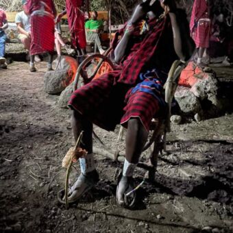 Safari Tanzania. Bezoek van gasten aan een Masai Boma. Een Masai zit op een zelf gefabriceerde stoel. Daarachter staan meerdere Masai mannende druk in gesprek zijn