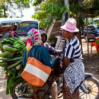 Reiservaring Tanzania. In Karatu staan 3 mensen bij elkaar. Een zit op een brommer met achterop grote trossen groene bananen