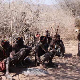 Tijdens een safari rondreis door Tanzania een activiteit. Gasten bezoeken de Hadzabe stam. Er zitten verschillende mannen op de grond in hun traditioneel kleding